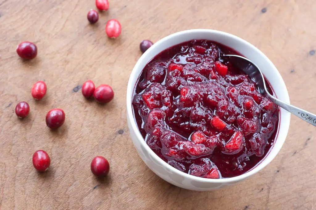 Top view of some chunky cranberry sauce in a bowl with a silver spoon and some fresh cranberries scattered about