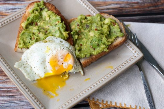 Avocado spread onto wheat pieces of toast with a half eaten over easy egg on a beige plate next to a fork and knife