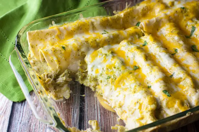 Closeup of clear casserole dish with a couple of enchiladas missing from the dish with a green napkin in the background