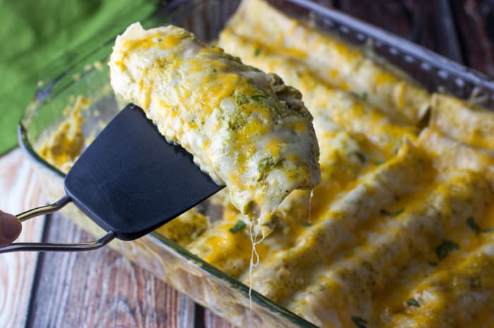 A black spatula taking out a single chicken enchilada from the casserole dish that's dripping with cheese