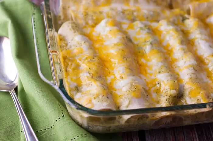 tortillas rolled up next to each other tightly in a casserole dish with melted cheese on top with a green napkin and large silver serving spoon