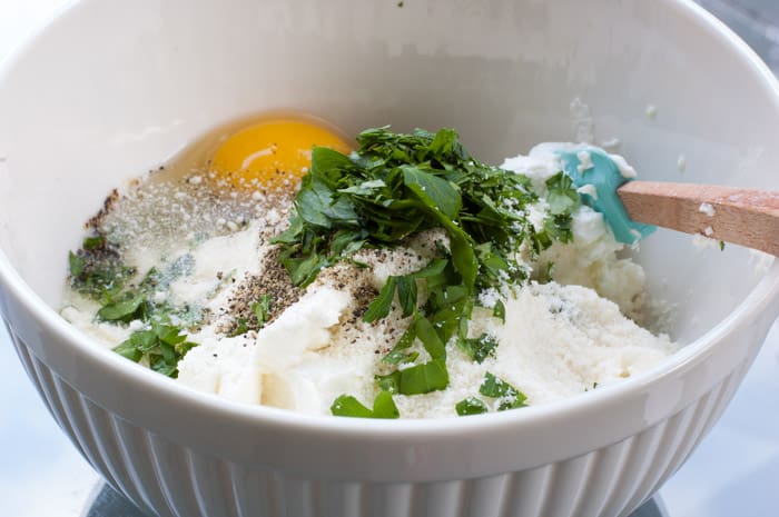 White ceramic bowl with ricotta cheese, egg, spices and parsley ready to be mixed with a spatula