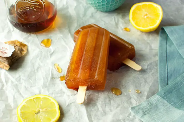 top view of two tea popsicles partially melted on parchment paper with lemon slices, tea bag, and jar of ice tea in the background