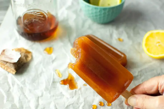 hand holding partially eaten tea popsicle with background showing ice tea bags, jar of ice tea and small teal bowl with lemon slices