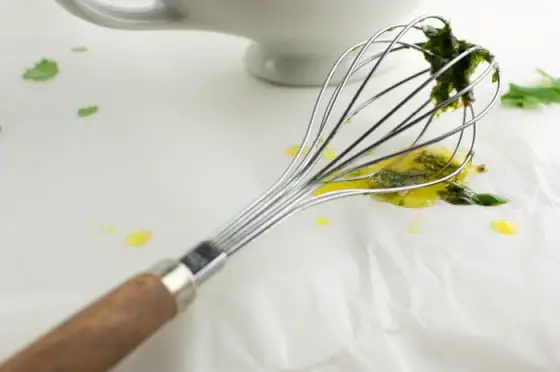 wooden and metal whisk with bits of honey lime cilantro sauce stuck to the end and drips of it all over parchment paper