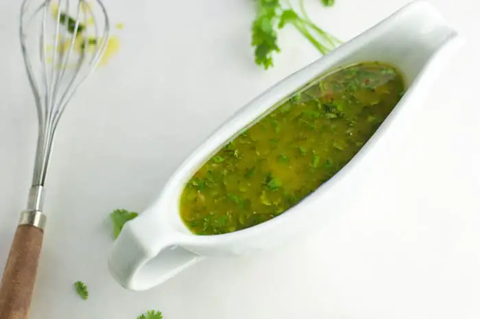 Top view of a white gravy dish filled with honey lime cilantro sauce with a dirty whisk sitting next to it and broken pieces of cilantro around it