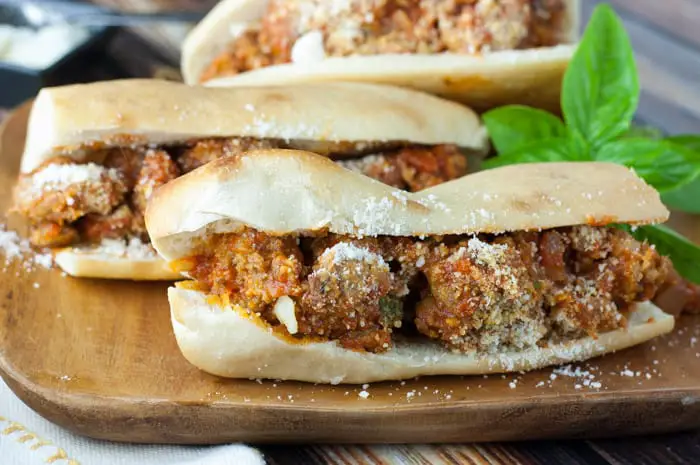 Wooden board with 3 long meatball sandwiches stacked together and a sprig of basil