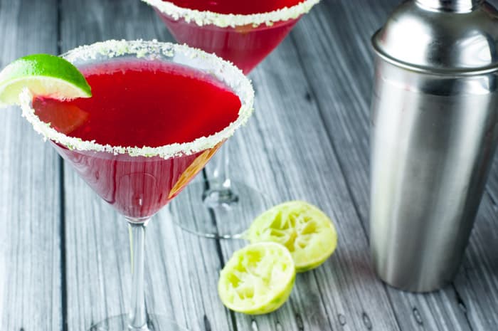 Shot from up above of 2 martini glasses filled with cape cod cosmopolitans with sugared rims and a slice of lime on the edge. 2 half used limes in the background with a silver cocktail shaker.