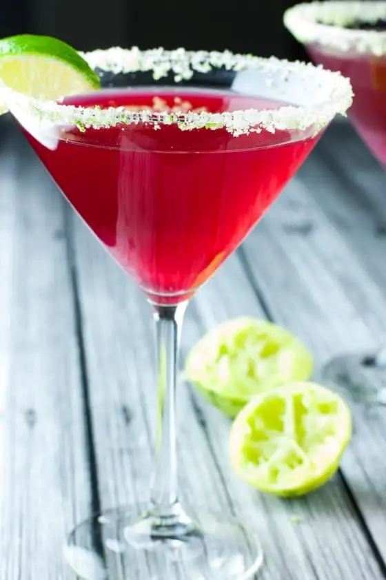 Side view of deep red cape cod cosmopolitan in a martini glass with the rim lined with green lime sugar with 2 half used limes in the background