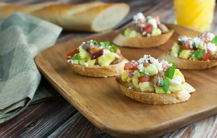 Small wooden cutting board that has 4 pieces of toasted baguette slices topped with breakfast toppings like eggs, sausage, and basil with a napkin and a glass of orange juice in the background