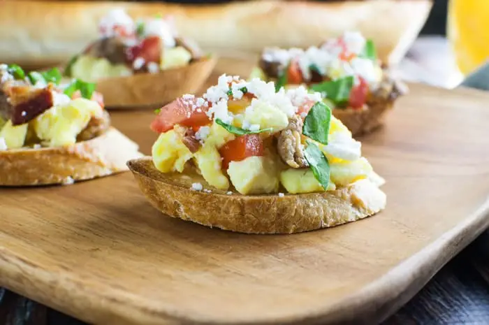 Side view of a toasted slice of baguette bread piled high with scrambled eggs, tomatoes, sausage, spinach, and feta