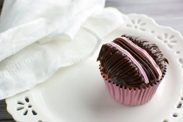 Cupcake in a pink wrapper with chocolate and strawberry frosting decorated with lines on a white plate with a white linen napkin