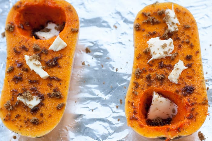 Two halves of butternut squash on tinfoil topped with cinnamon, brown sugar, and pats of butter