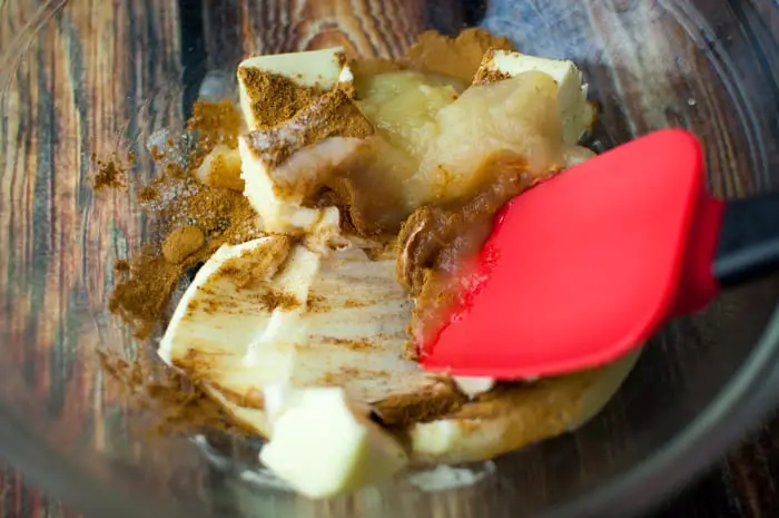 Clear glass bowl with softened butter, apple sauce, and apple pie spices being mixed together with red spatula 