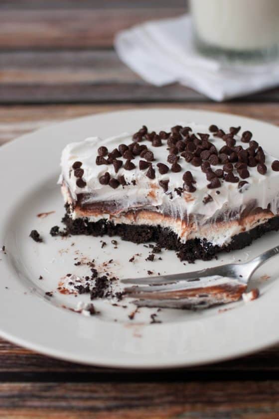 thick layers of cookie crust, cream cheese, chocolate pudding, whip cream and chocolate chips on top half eaten on a plate with a glass of milk