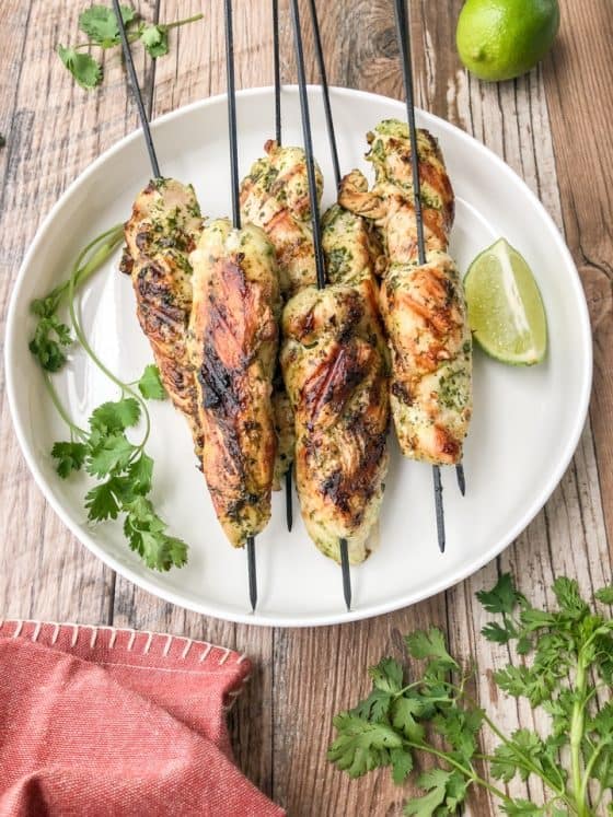 Five chicken skewers on a white plate with lime and cilantro
