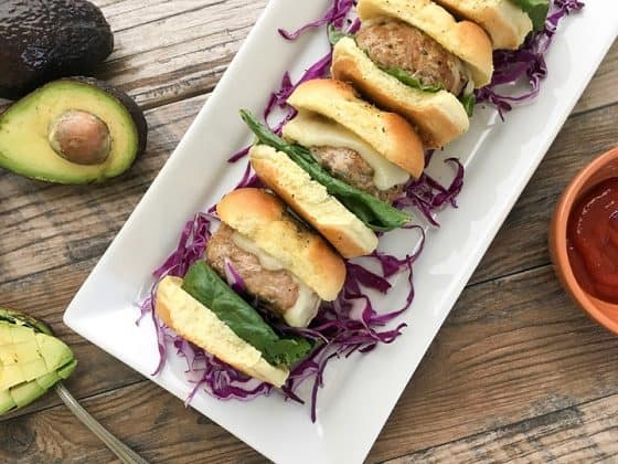 Four turkey burger sliders stacked up on a rectangle white plate with an halved avocado in the background