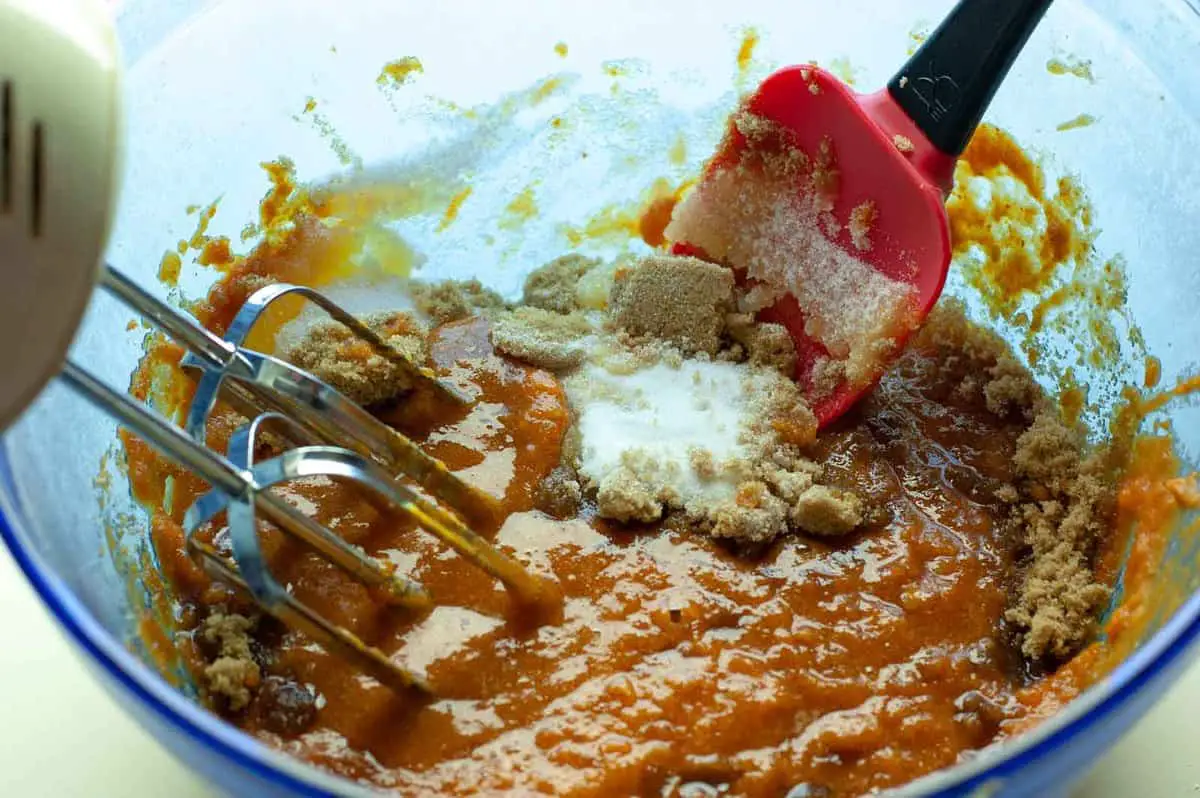 Electric beater and red spatula mixing together canned pumpkin with brown and white sugar