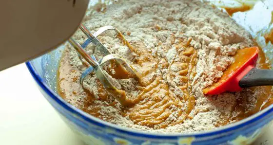 Electric beater and red spatula mixing together pumpkin mix with flour in a blue bowl