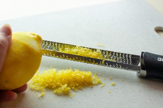 Citrus zester holding a pile of lemon zest with a partially zested lemon on the side