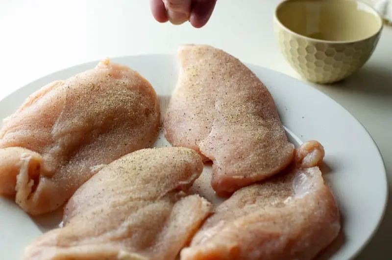 Four raw chicken breasts on a white plate being seasoned with salt and pepper