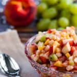 Chopped fruit salad in a crystal bowl with peaches and green grapes in the background