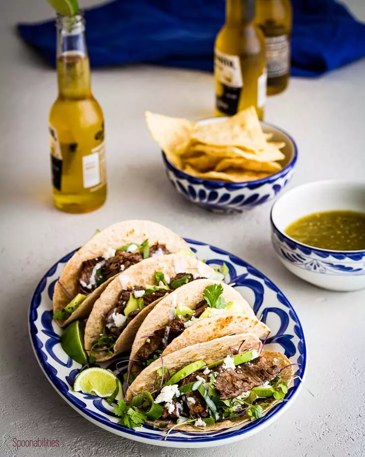 Plate with 4 steak tacos with cilantro and lime wedges and some Corona beers in the background