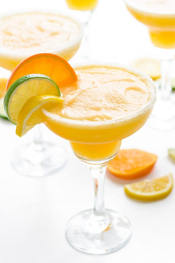 Bright orange frozen margarita in a glass with slices of lemon, lime, and orange on the rim.
